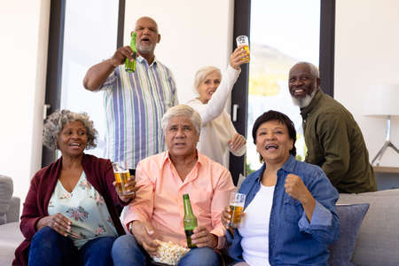 Multiracial excited senior friends with beer and popcorns enjoying match in nursing home. Sport, curiosity, alcohol, food, unaltered, togetherness, support, assisted living and retirement concept.の写真素材