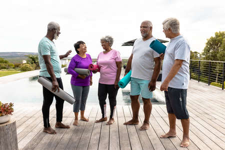 Happy multiracial senior friends holding exercise mats talking while standing on hardwood floor. Yard, copy space, unaltered, togetherness, support, assisted living, retirement home, fitness.の写真素材