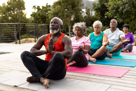 Multiracial senior friends meditating while sitting on mats against plants in yard at nursing home....