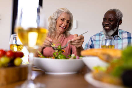 Low angle view of multiracial cheerful senior couple having lunch at dining table in nursing home. Wine, meal, laughing, love, unaltered, togetherness, support, assisted living and retirement.の写真素材
