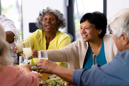 Multiracial cheerful senior friends toasting drinks while having lunch at dining table. Nursing...