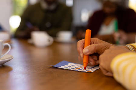 Cropped hands of biracial senior woman marking off numbers on bingo card over table in nursing home. Felt-tip pen, luck, leisure game, unaltered, support, assisted living, retirement concept.の写真素材