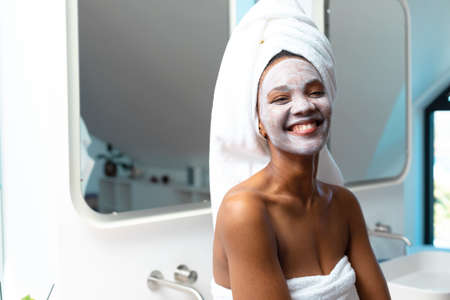 Portrait of cheerful young african american woman with face cream on face in bathroom, copy space. Unaltered, spa, home, relaxation, pampering, detox, body care and wellbeing concept.の写真素材