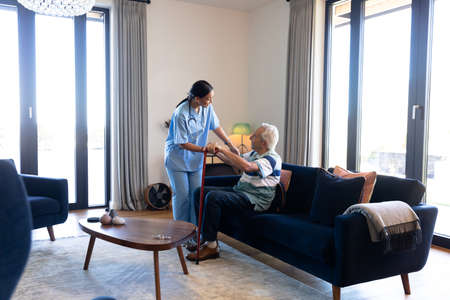 Biracial female health worker helping caucasian senior man to get up from the couch at home. Medical care and retirement senior lifestyle conceptの写真素材