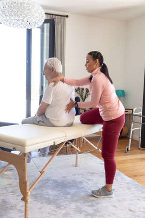 Biracial female physiotherapist giving back massage therapy to caucasian senior man at home....