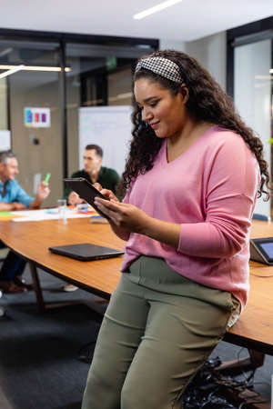Smiling young biracial businesswoman using tablet pc while colleagues in background. Unaltered,...