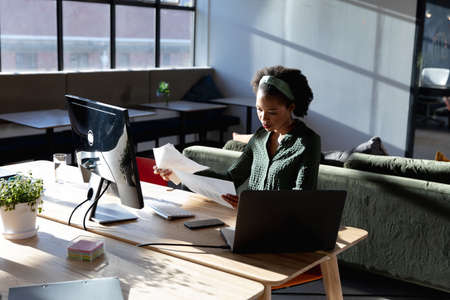 African american young businesswoman analyzing documents while sitting at computer desk in office. Unaltered, creative business, workplace, technology, occupation, paperwork.の写真素材