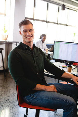 Portrait of smiling young caucasian businessman with computer at desk in office. Unaltered, creative...