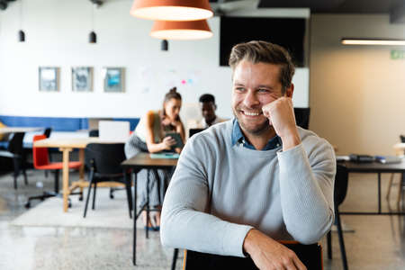 Smiling caucasian mid adult businessman sitting on chair in creative office. Unaltered, creative business, workplace, occupation, positive emotion.の写真素材