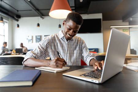 Smiling young african american businessman writing in diary and using laptop in creative office....