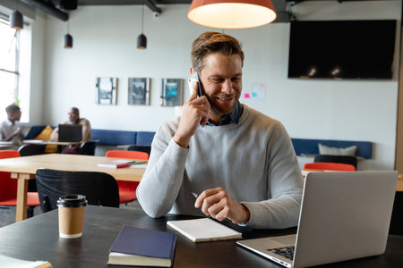 Smiling caucasian mid adult businessman talking on smart phone in creative office. Unaltered,...