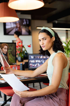 Portrait of confident biracial young businesswoman with photographs at desk in office. Unaltered,...