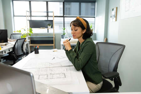Asian young female architect with blueprint at table in office. Unaltered, workplace, creative business, design professional, floor plan.の写真素材