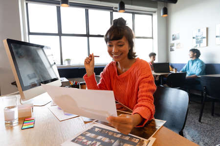 Smiling creative asian businesswoman with photograph at computer desk in office. Unaltered, creative...