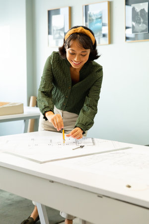 Smiling asian young female architect drawing blueprint at table in creative office. Unaltered, workplace, creative business, design professional, floor plan.の写真素材