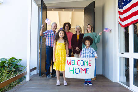Multiracial multigeneration family with welcome home text and flags of america standing at entrance. Unaltered, family, love, togetherness, childhood, happy, greeting, patriotism, house, retirement.の写真素材