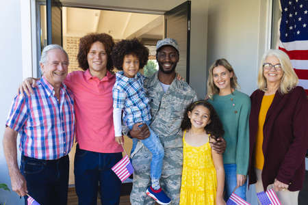 Portrait of smiling multiracial soldier with multigeneration family standing at entrance of house. Flag of america, homecoming, unaltered, family, togetherness, childhood, military, patriotism.の写真素材