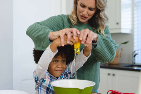 Multiracial mother making face while assisting son with afro hair in breaking egg in bowl in...