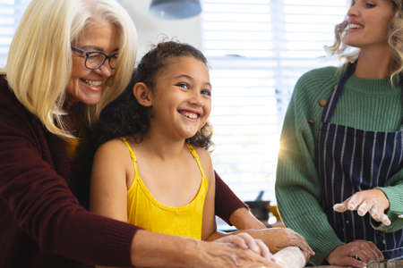 Happy woman looking at grandmother assisting granddaughter in rolling dough on table in kitchen....