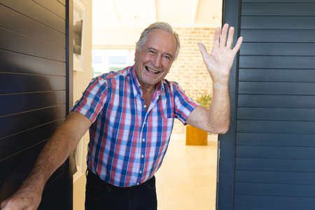 Portrait of cheerful caucasian senior man waving hand and welcoming while standing at entrance....