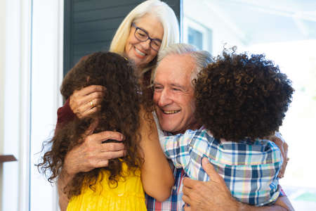 Multiracial grandchildren embracing cheerful grandparents and welcoming them at home at entrance....