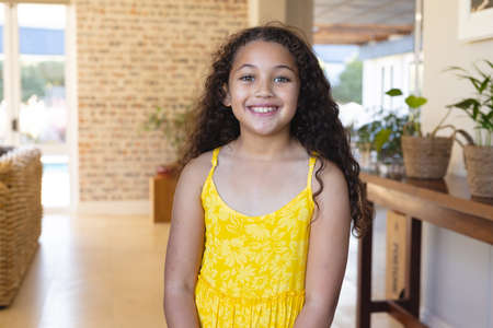 Portrait of smiling biracial cute girl with long curly hair wearing yellow dress standing at home....