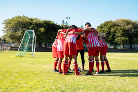 Male multiracial players in red uniform with arms around discussing and huddling at playground....