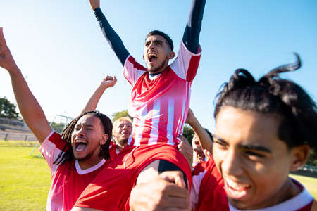 Multiracial male players carrying cheerful teammate on shoulders screaming while celebrating goal. Match, playground, unaltered, soccer, sport, teamwork, togetherness, competition and winning.の写真素材