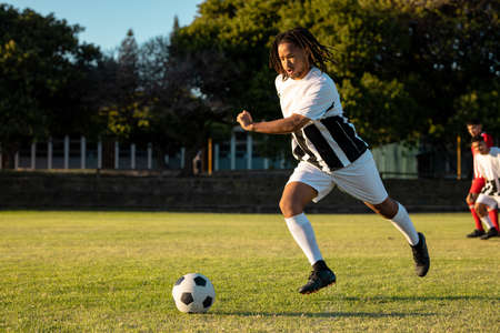 Biracial male player with dreadlocks running and kicking ball during match on grassy playground. Unaltered, soccer, sport, team sport, effort, summer and competition concept.の写真素材