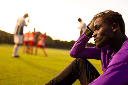 African american stressed male goalkeeper with head in hand sitting on field against clear sky. Copy space, playground, frustration, loss, sadness, unaltered, soccer, competition and sport concept.の写真素材