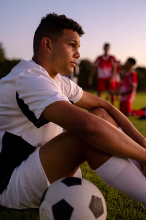 Side view of thoughtful sad caucasian male player looking away and sitting against clear sky. Sunset, playground, frustration, loss, stress, contemplation, unaltered, soccer, competition, sport.の写真素材