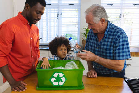 Multiracial grandfather and father teaching boy to recycle waste by throwing plastic bottles in bin. Multigeneration, unaltered, together, childhood, unaltered, environmental conservation, garbage.の写真素材