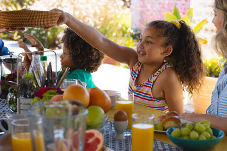 Multiracial cute girl passing food while having breakfast with mother and brother at dining table....