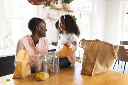 Image of happy african american mother and daughter unpacking shopping bags in kitchen. Family and...