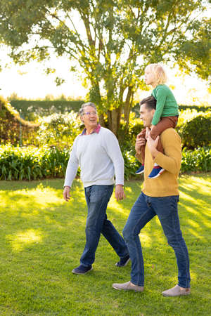 Vertical image of happy three generation caucasian men walking in garden. Family and spending...