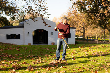 Image of happy caucasian senior man swiping leaves in garden. Lifestyle, autumn, spending time at home and garden concept.の写真素材