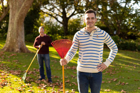 Image of caucasian father and adult son swiping leaves in garden. Family and spending quality time together concept.の写真素材