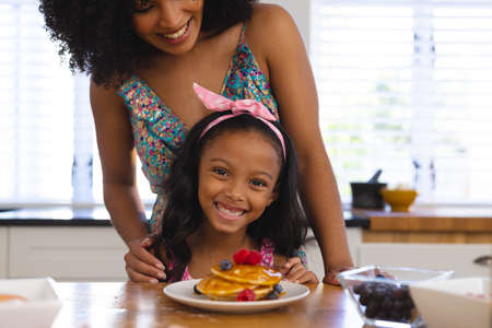 Midsection of happy biracial mother and daughter with tempting pancakes on table at home. Portrait, unaltered, family, togetherness, childhood, food, breakfast and lifestyle concept.の写真素材
