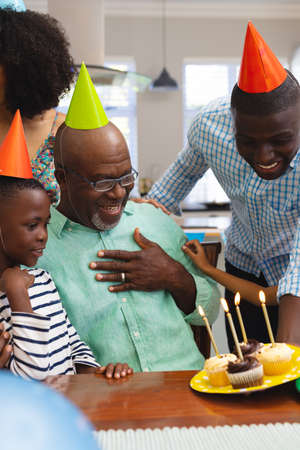 Multiracial senior man wearing party hat looking at cupcakes while celebrating birthday with family. Happy, unaltered, togetherness, sweet food, retirement, enjoyment and home concept.の写真素材