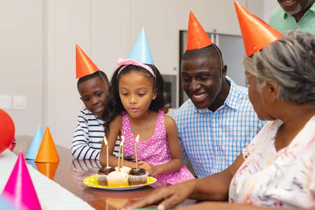Multiracial girl wearing party hat blowing candles while celebrating birthday with family at home. Unaltered, togetherness, love, childhood, cupcake, enjoyment and retirement.の写真素材
