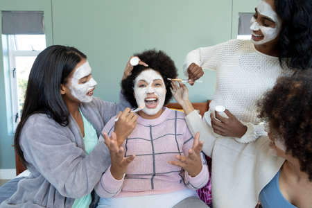 Biracial young females applying beauty cream on cheerful friend's face with afro hair screaming. Skin care, pampering, unaltered, friendship, togetherness, lifestyle and home concept.の写真素材