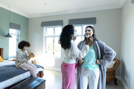 Biracial young woman applying beauty cream on smiling friend's face and female sitting on bed....