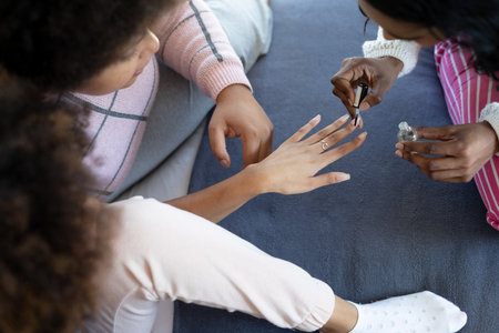 High angle view of biracial young woman painting fingernails of female friend on bed at home. Body care, pampering, beauty product, unaltered, friendship, togetherness, lifestyle and home.の写真素材