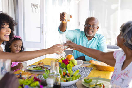 Multiracial senior man raising toast while having lunch with multigeneration family at dining table. Home, happy, drink, unaltered, togetherness, love, childhood and retirement concept.の写真素材