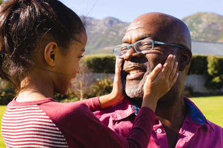 Biracial granddaughter touching smiling african american bald grandfather's face in summer....