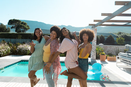 Carefree biracial female friends posing while hanging out at poolside against clear sky, copy space. Summer, unaltered, friendship, togetherness, social gathering, enjoyment and weekend activities.の写真素材