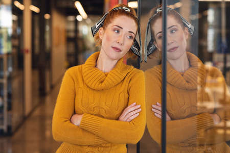 Thoughtful mid adult caucasian businesswoman with arms crossed leaning on glass wall in office....