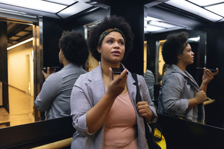 Mid adult biracial businesswoman talking on smartphone speaker while standing in elevator....