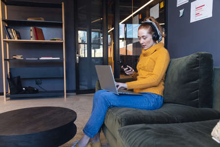 Mid adult caucasian businesswoman using smart phone and laptop while sitting on sofa in office....