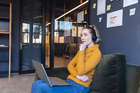 Thoughtful mid adult caucasian businesswoman with laptop sitting on sofa in office. Unaltered,...